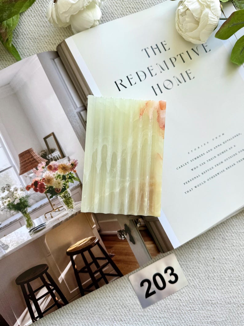 May include: A rectangular, light green and orange-toned soap bar with vertical ridges. The soap is displayed on an open book titled "THE REDEMPTIVE HOME". A small metal tag with the number "203" is visible.