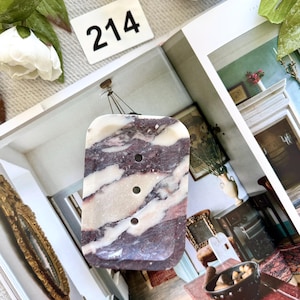 May include: A rectangular pendant with a marbled pattern of burgundy and white. The pendant has three circular holes and is suspended by a thin cord. A white tag with the number "214" is visible in the background.