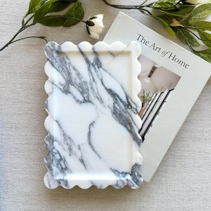 May include: A rectangular marble tray featuring a scalloped edge design. The tray has a white base with prominent grey veining throughout the surface. The tray is positioned next to a book titled The Art of Home and a stem of white flowers.