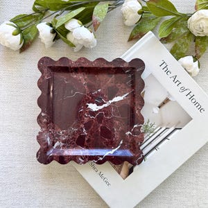 May include: A square, burgundy marble tray with a scalloped edge. The tray has white veining and is next to a book titled "The Art of Home" and white flowers.