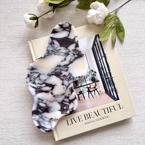 May include: A marbled massage tool in white, cream, and burgundy sits on a book titled "LIVE BEAUTIFUL." The tool is next to a sprig of white flowers and green leaves. The book cover features a photograph of a room.