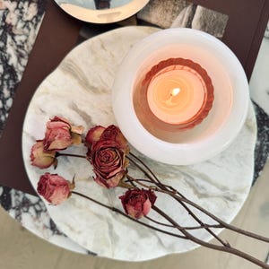 May include: A white marble circular tray holding a lit candle in a white glass holder with a fluted interior and several dried red roses on thin brown stems. A dark brown book with the text AMBER LE is partially visible beneath the tray.