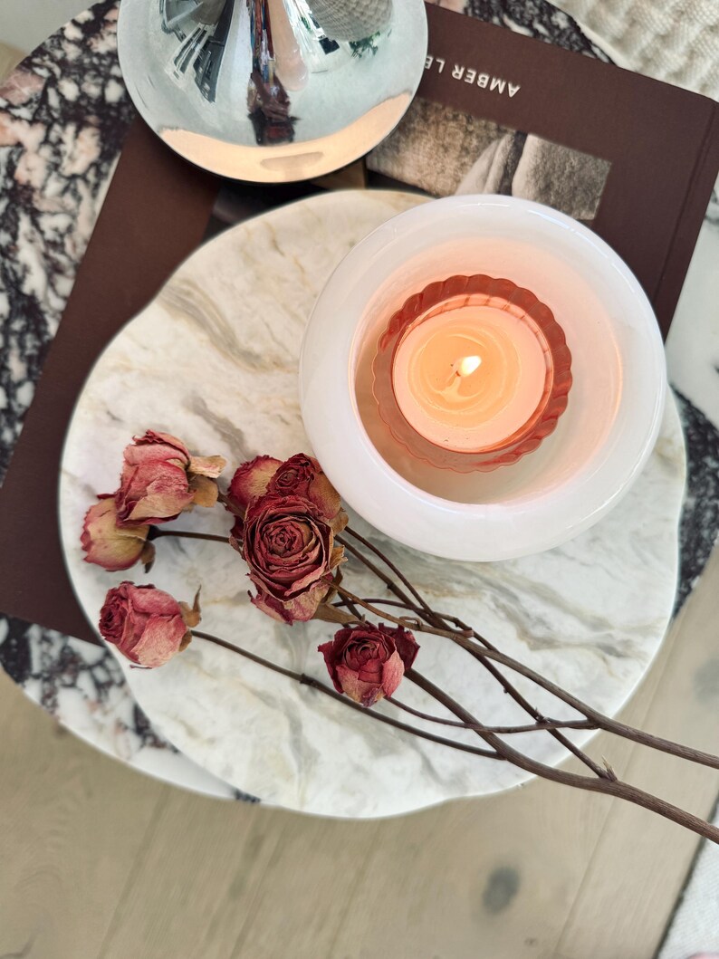 May include: A white marble circular tray holding a lit candle in a white glass holder with a fluted interior and several dried red roses on thin brown stems. A dark brown book with the text AMBER LE is partially visible beneath the tray.