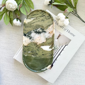 May include: An oval-shaped decorative tray with a marbled green and white pattern. The tray is resting on a book titled "The Art of Home" and is surrounded by white flowers and green leaves.