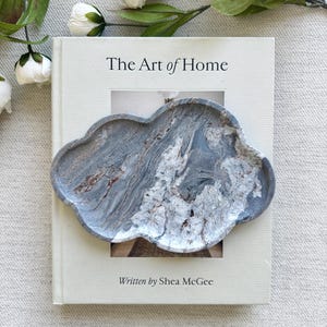 Może przedstawiać: Taca w kształcie chmury z szaro-białym wzorem marmurkowym, umieszczona na twardej oprawie książki zatytułowanej The Art of Home autorstwa Shea McGee. Taca posiada wirujące żyłki w kolorze ciemnoszarym, jasnoszarym i białym na całej powierzchni.