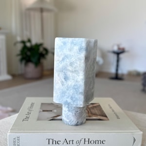 May include: A light blue, rectangular decorative object with a square top and a round base. The object is sitting on a book titled "The Art of Home". The book is white with black text. The object has a marbled appearance.