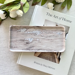 Puede incluir: Una bandeja decorativa rectangular con diseño de mármol en tonos beige, marrón y blanco. La bandeja está sobre un libro titulado "The Art of Home" de Shea McGee, con flores blancas en el fondo.