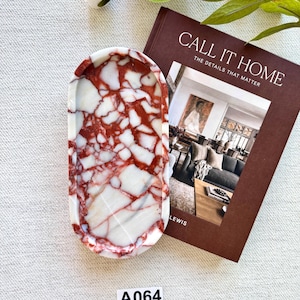Könnte beinhalten: Ovaler Marmortablett mit rotem und weißem Muster, neben einem Buch mit dem Titel "CALL IT HOME". Das Tablett hat eine polierte Oberfläche und eignet sich zur Präsentation kleiner Gegenstände. Ein weißes Etikett mit "A064" ist sichtbar.