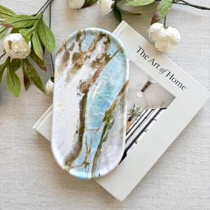 May include: An oval-shaped tray featuring a marbled pattern with swirls of light blue, white, and earthy brown tones. The tray has a smooth, polished surface with a distinct veined appearance, showcasing a natural stone-like aesthetic.