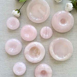 Peut inclure: Une collection de neuf bols et objets circulaires en pierre translucide rose pâle, lisses et de tailles variées. Chaque pièce présente une surface polie et arrondie avec une indentation centrale, révélant des nuances de rose laiteux naturel.