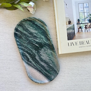 Puede incluir: Una bandeja oblonga de mármol verde y blanco. La bandeja tiene un acabado pulido y un diseño veteado natural. Un libro con las palabras "LIVE BEAUTIFUL" está al lado de la bandeja.