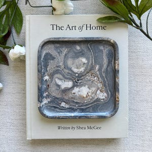 Peut inclure: Un plateau carré aux coins arrondis présentant un motif marbré dans des tons gris anthracite, ardoise et blanc avec des inclusions cristallines texturées. Le plateau est posé sur un livre à couverture rigide blanc intitulé The Art of Home par Shea McGee.