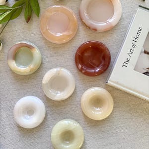 Op de afbeelding: Een assortiment van acht gepolijste stenen onderzetters in verschillende kleuren, waaronder wit, roze, bruin en groen. De onderzetters zijn cirkelvormig met een verzonken midden. Een boek getiteld "The Art of Home" is zichtbaar.
