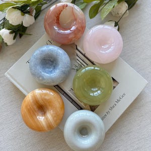 May include: Six donut-shaped stone carvings in shades of pink, blue, green, orange, and white. The carvings are arranged on a book with the text "written by Shea McGee". White flowers and green leaves are in the background.