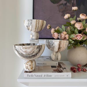 May include: Three decorative bowls crafted from natural stone with swirling patterns of beige, brown, and cream. Each bowl features a wide, rounded basin supported by a spherical base. The stone displays unique, organic veining throughout.