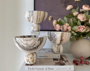 May include: Three decorative bowls crafted from natural stone with swirling patterns of beige, brown, and cream. Each bowl features a wide, rounded basin supported by a spherical base. The stone displays unique, organic veining throughout.