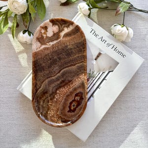 May include: An oval-shaped tray crafted from polished brown onyx stone, featuring natural banded patterns in shades of dark chocolate, tan, and cream. The tray has a smooth, glossy finish with a raised edge and a distinct, dark circular inclusion.