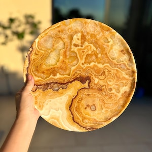 May include: A circular tray crafted from polished natural stone, featuring a swirling pattern of warm amber, golden yellow, and deep brown tones. The surface displays translucent crystalline textures and organic banding throughout the entire round piece.