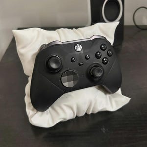May include: A black wireless video game controller featuring a multi-directional pad with a faceted metallic finish, two analogue sticks, four primary action buttons, and a central Xbox logo button, resting against a small, white, gathered fabric pillow.