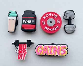 May include: Set of six colorful shoe charms with a gym theme. The charms include a protein shaker, a protein powder container, a weight plate, a dumbbell, a barbell with the text "No Days Off", and the word "GAINS" in pink and yellow.