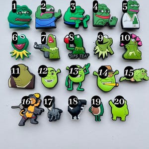 May include: Twenty colorful cartoon character shoe charms, including a frog drinking tea, a frog with a gun, a frog with hearts, a Shrek character with a cigar, and a cat in a knight costume.