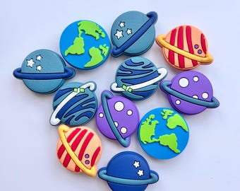 May include: A collection of twelve circular shoe charms featuring space-themed designs, including planets with rings, Earth with green continents on a blue background, and various celestial bodies in shades of blue, purple, orange, and yellow.
