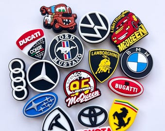 May include: A collection of colorful shoe charms featuring various automotive brand logos and character designs, including Ducati, Ford Mustang, Volkswagen, Lamborghini, BMW, Audi, Mercedes-Benz, Lightning McQueen, Bugatti, Subaru, Tesla, Toyota, and Ferrari.