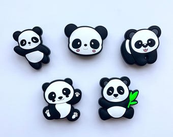May include: A set of five black and white panda-shaped charms. The designs include a panda waving, a panda face, a sitting panda, a panda with raised paws, and a panda holding a bright green bamboo stalk. Each piece features a smooth finish.