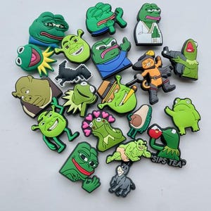 Könnte beinhalten: Eine Sammlung von 18 grünen Frosch- und Meme-Charakter-Schuh-Anhängern. Die Anhänger sind aus Gummi und zeigen verschiedene Ausdrücke und Posen. Einige der Anhänger enthalten den Text "Sips Tea" und eine Schwarzweißillustration eines Dinosauriers.