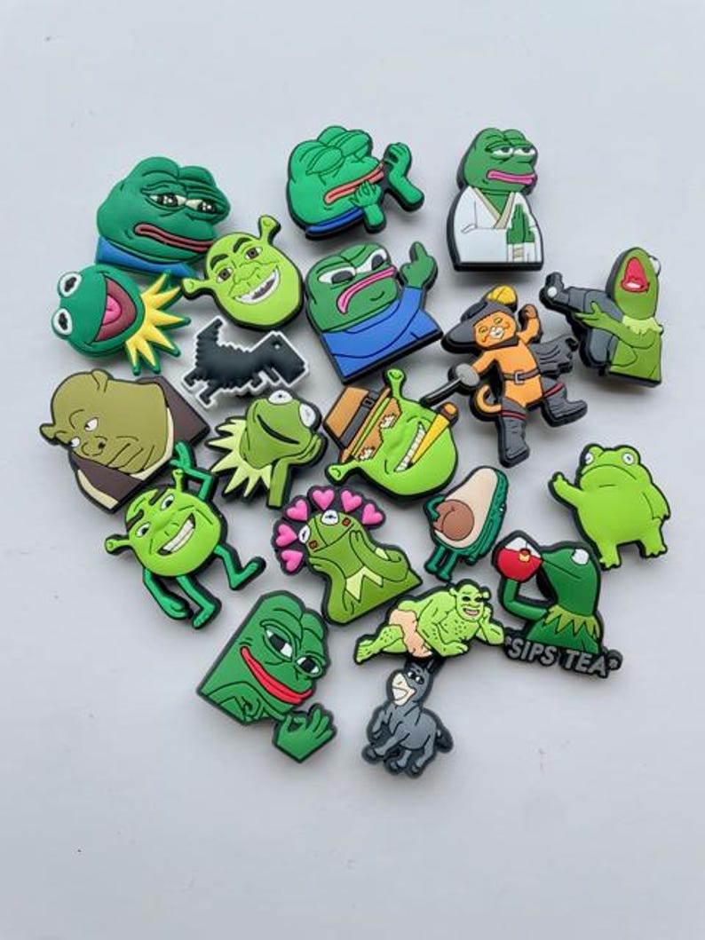 May include: A collection of 18 green frog and meme character shoe charms. The charms are made of rubber and feature various expressions and poses. Some of the charms include the text "Sips Tea" and a black and white illustration of a dinosaur.