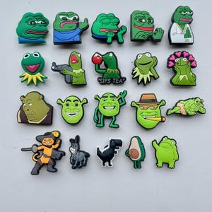 May include: A collection of 20 green frog and Shrek themed shoe charms. The charms feature various expressions and poses, including a frog sipping tea, a frog with a gun, and Shrek with a cigar.