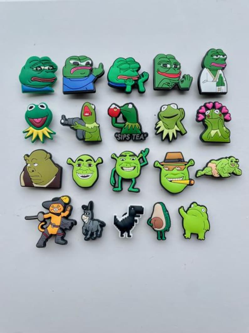 May include: A collection of 20 green frog and Shrek themed shoe charms. The charms feature various expressions and poses, including a frog sipping tea, a frog with a gun, and Shrek with a cigar.