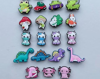 May include: A collection of colorful shoe charms featuring various characters including frogs, cows, dinosaurs, axolotls, a llama, a rabbit, and mushrooms. The set includes diverse designs such as a frog with a guitar, a frog in a car, and a frog with a knife.