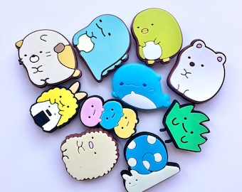 May include: A collection of eleven colorful silicone shoe charms featuring various cartoon characters including a blue dinosaur, a green penguin, a white bear, a yellow rice ball, a blue whale, a green weed, a snail, and several small round faces.
