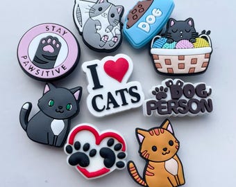 May include: A collection of nine colorful shoe charms featuring various cat and dog designs, including text that reads STAY PAWSITIVE, I LOVE CATS, and DOG PERSON, alongside illustrations of cats, a dog bowl, a yarn basket, and paw prints.