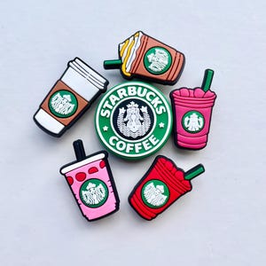Könnte beinhalten: Eine Sammlung farbenfroher Starbucks-Charms. Die Charms umfassen Kaffeetassen, Frappuccinos und ein rundes Logo mit dem Starbucks-Namen und -Logo. Die Charms sind in Braun-, Rosa- und Rottönen gehalten, mit grünen und weißen Akzenten.