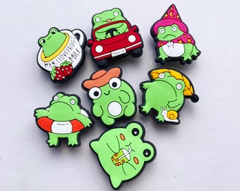 May include: A collection of seven colorful frog-themed charms. Each charm features a green frog in various poses and outfits, including a car, a witch hat, and a life preserver. Some charms have text, such as "Man, I love frogs, mile".