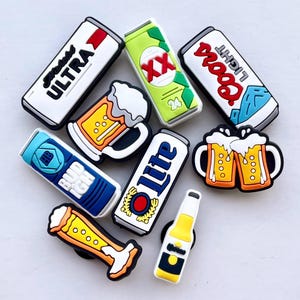 May include: A collection of colorful shoe charms featuring beer cans, beer mugs, and beer bottles. The charms include the logos of various beer brands, with a variety of colors including white, green, blue, and yellow.