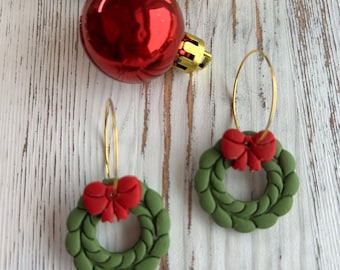 May include: A pair of gold hoop earrings featuring green wreath charms with red bows. Christmas ornaments in red, green, and gold are in the background, creating a festive holiday theme.