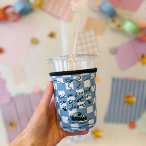 Puede incluir: Un vaso de plástico transparente con pajita y funda a cuadros azules y blancos. La funda tiene el texto "tis so sweet to trust in Jesus" y una ilustración de pastel. El vaso está sostenido en una mano.