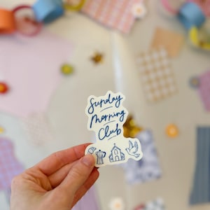 May include: A white sticker with the text "Sunday Morning Club" in a handwritten font. The sticker also features illustrations of a coffee pot, church, and dove. The background includes colorful paper chains and patterned paper cutouts.