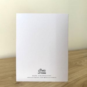 May include: A white greeting card with the text "Shea Lettering" in black script. Below the name is the text "MADE IN KANSAS CITY" and the website address. The card is on a wooden surface.
