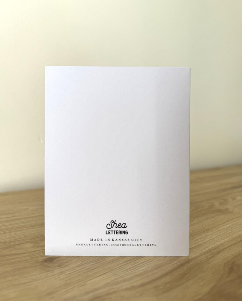 May include: A white greeting card with the text "Shea Lettering" in black script. Below the name is the text "MADE IN KANSAS CITY" and the website address. The card is on a wooden surface.