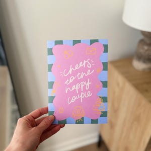 May include: A greeting card with a pink blob design and the words "Cheers to the happy couple" in white script. The card has a blue and green checkered background and small yellow illustrations.