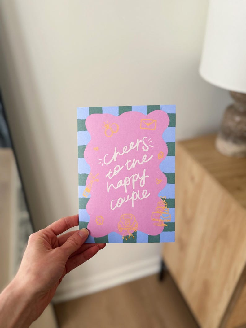 May include: A greeting card with a pink blob design and the words "Cheers to the happy couple" in white script. The card has a blue and green checkered background and small yellow illustrations.