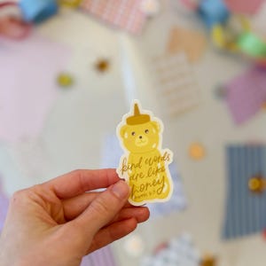 May include: A yellow sticker in the shape of a bear holding a honey jar. The sticker has the text "kind words are like honey" and "Proverbs 16:24". The sticker is held in a hand.