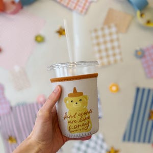 May include: A clear plastic tumbler with a straw, held by a hand. The tumbler has a beige sleeve with a cartoon bear design and the text "kind words are like honey". The background includes colourful paper decorations.
