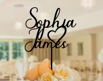 May include: A black cake topper with the names "Sophia" and "James" in elegant script, with a heart-shaped design between the names. The topper is placed on a cake decorated with yellow and white flowers.