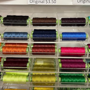 May include: A display of colorful spools of thread, arranged in clear acrylic shelves. The thread colors include shades of brown, black, teal, blue, orange, red, burgundy, pink, purple, yellow, and green. The spools are neatly organized, showcasing a variety of sewing supplies.