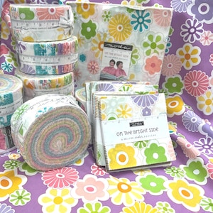 May include: A collection of fabric rolls and fat quarters featuring a floral print with a retro 70s vibe. The fabric is white with colourful flowers and the text "On the Bright Side" by Moda Sister Designs.